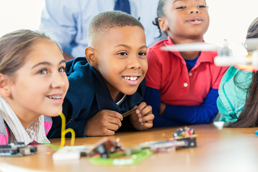Elementary age Hispanic and African American little boys and little girls are students in public elementary school STEM program. Students are learning about science, technology, engineering, and math while studying a drone. Children are smiling and wearing casual clothing.